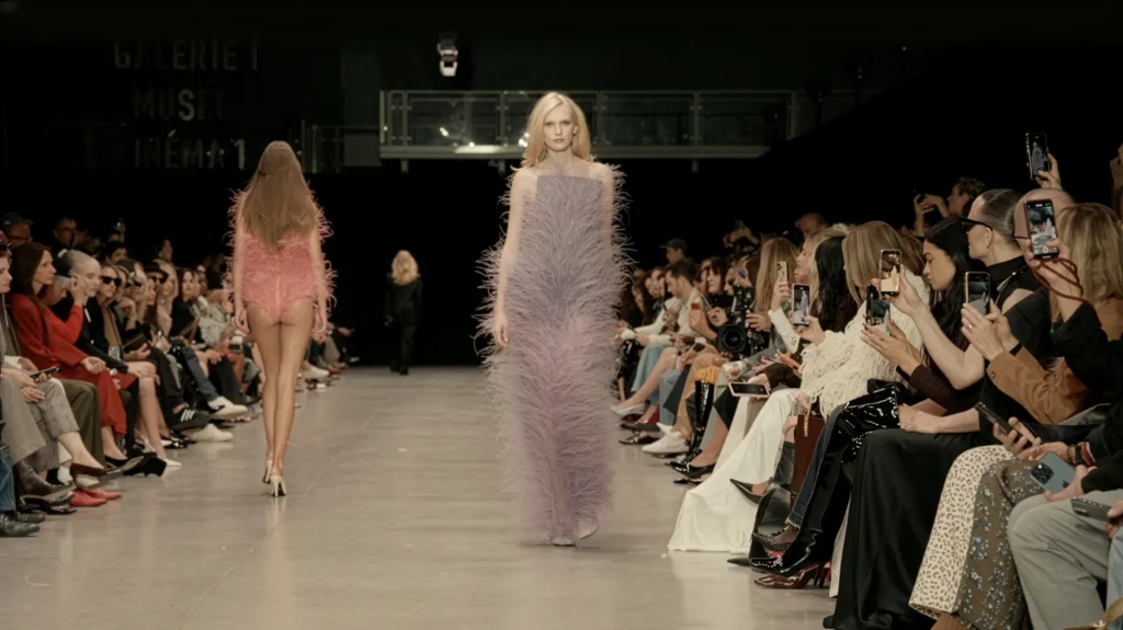 Models on the runway at Paris Fashion Week wearing Stella McCartney designs made with Fevvers’ plant-based feather alternative, in pink and lavender looks, as the audience photographs the show.